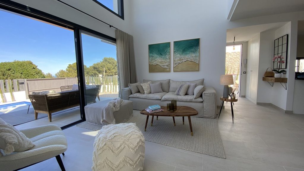 Salón de doble altura en Colònia de Sant Pere, Mallorca, con sofá beige, cuadros de mar y vistas a la piscina