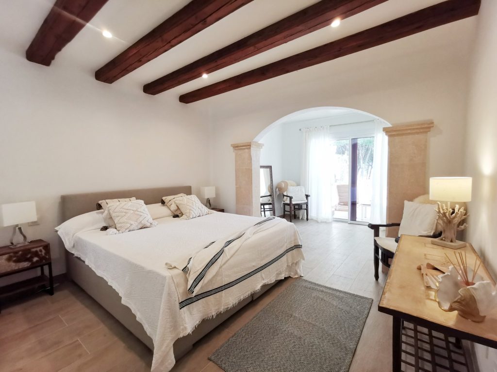 Dormitorio reformado en Santa Ponsa, Mallorca, con vigas de madera, arco de piedra y ropa de cama blanca