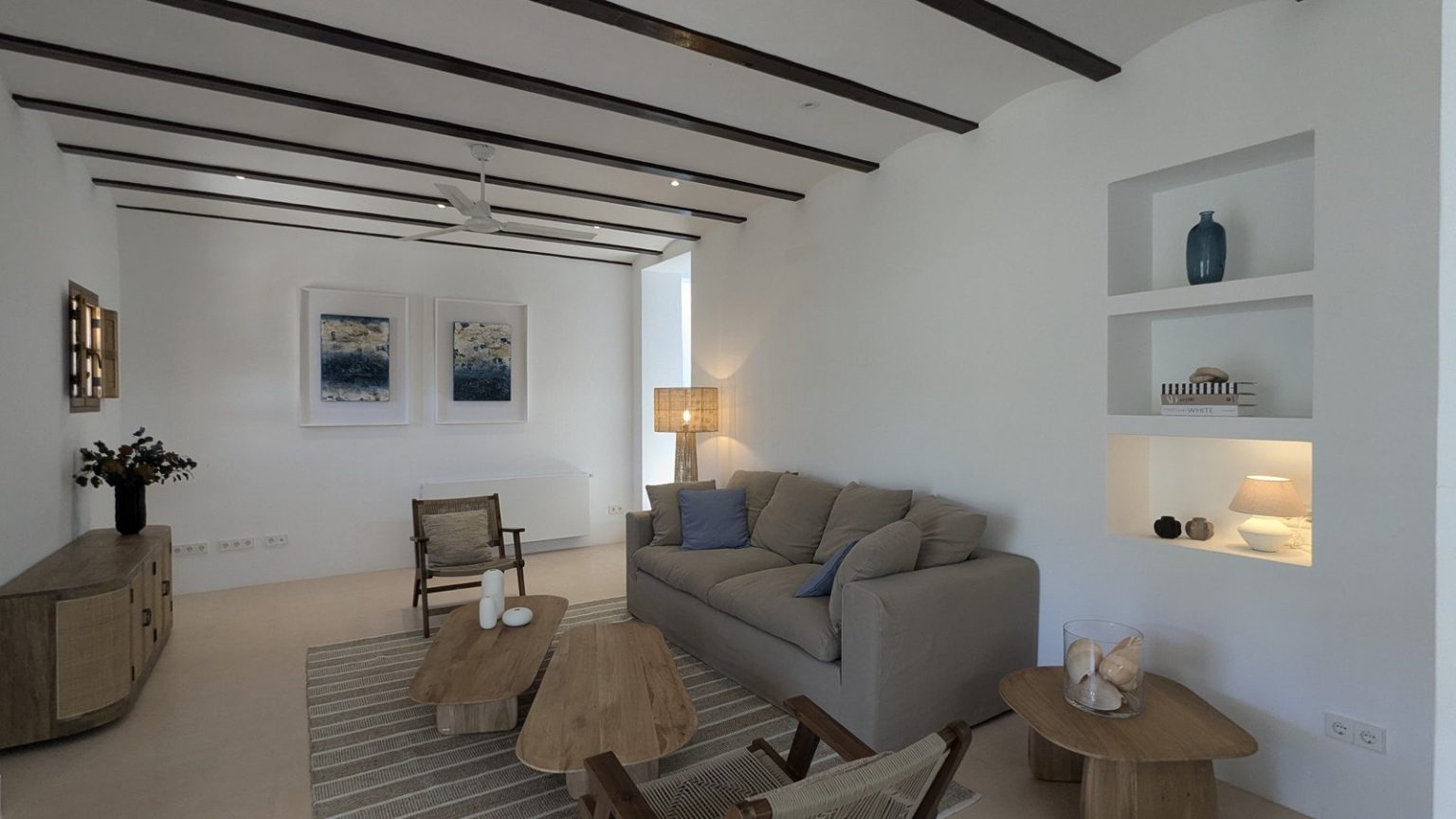 Salón blanco con vigas vistas, sofá gris y hornacinas decoradas en una finca de Ibiza
