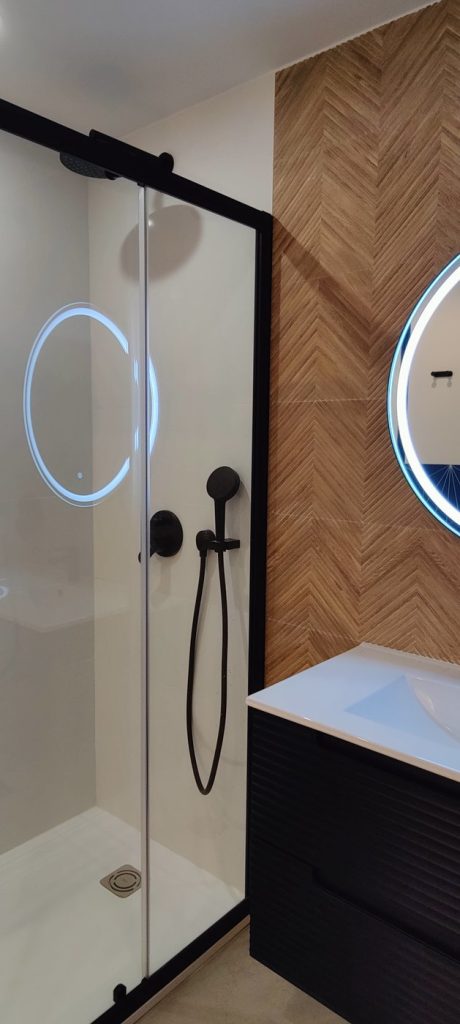 Baño reformado en Madrid con ducha de perfilería negra, pared en espiga de madera y espejo retroiluminado